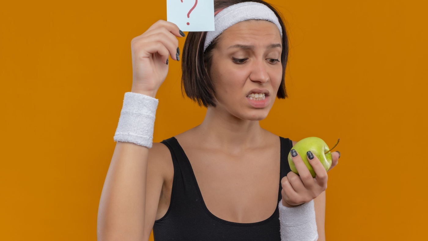 young fitness girl in sportswear with headband showing reminder paper with question mark holding green apple looking at it confused and very anxious standing over orange background